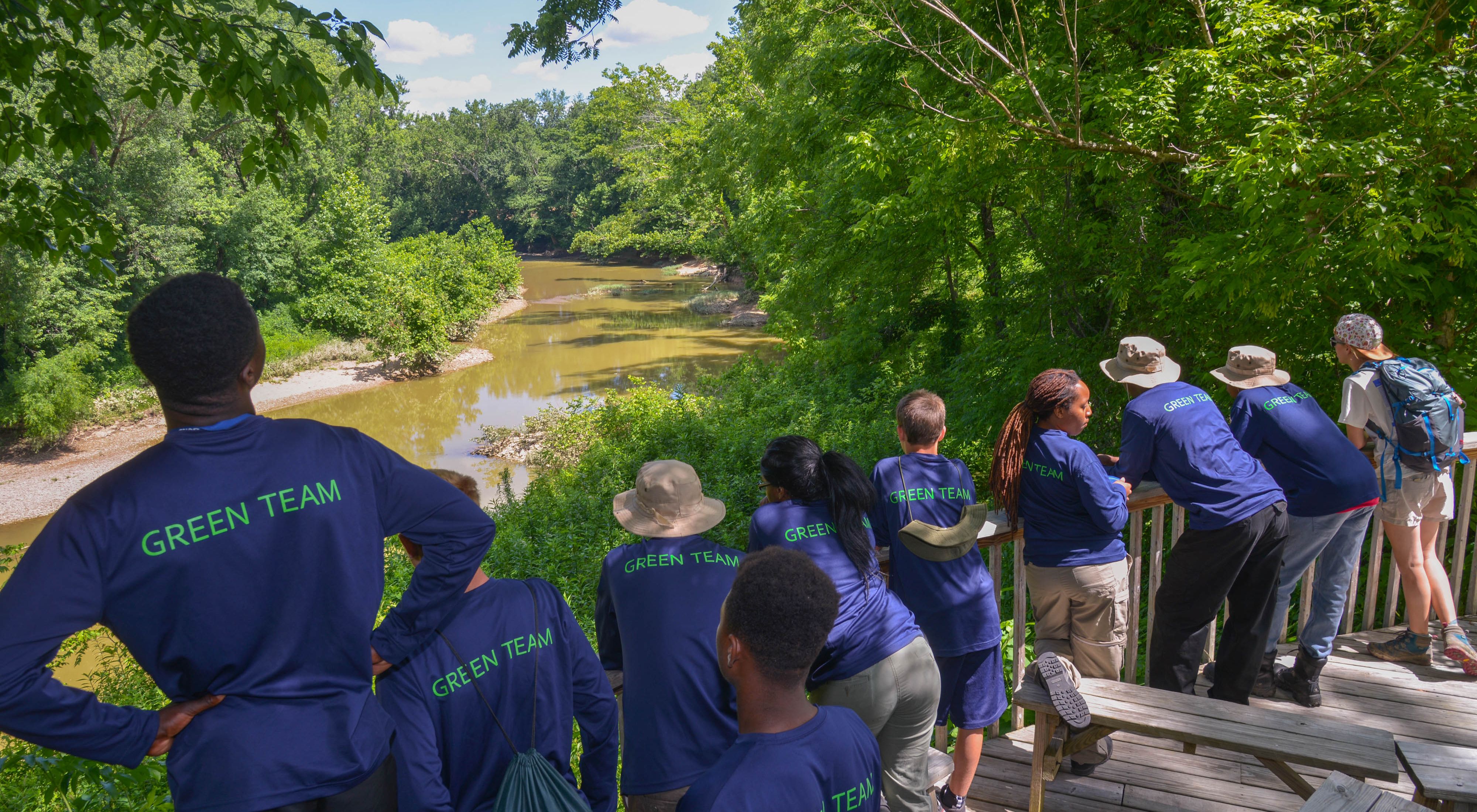 A row of teenagers in matching blue shirts stand along a wooden rail overlooking a river, surrounded by lush, green leaves.