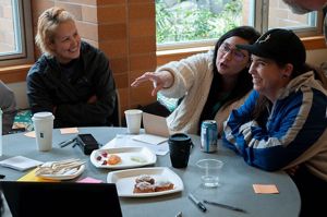 Three people sit together at a table talking and smiling. one is pointing at something across the table.