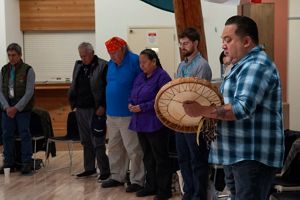 A group of people standing in a circle with their heads bent and eyes closed. One person holds a drum.