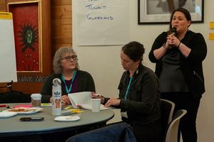 a person standing speaking into a microphone while two others sit at a table.