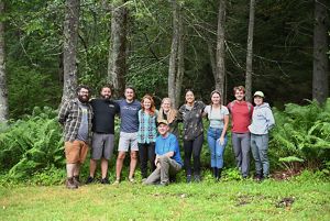 A group of 10 people pose together arm in arm at the edge of a forest on Warm Springs Mountain.