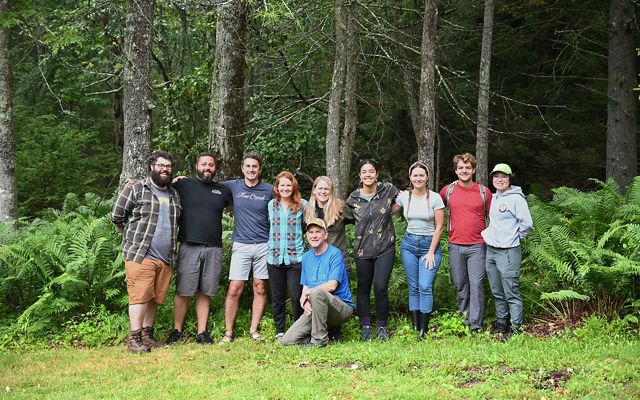 A group of 10 people pose together arm in arm at the edge of a forest on Warm Springs Mountain.