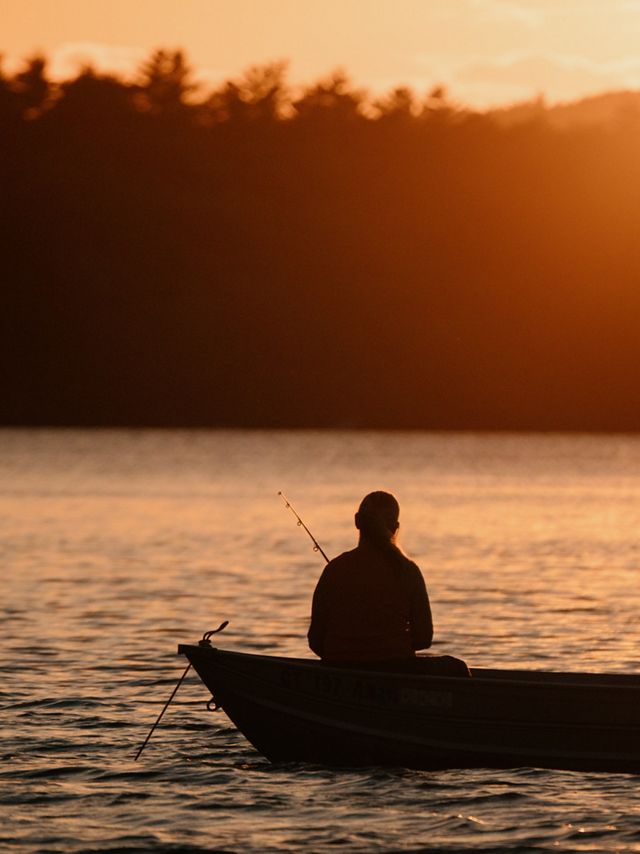 A silhouetted person sits with her back to the camera in a small boat holding a fishing rod in a body of water. 