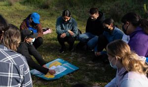 Group of students sitting in a circle in the grass looking down at a map.