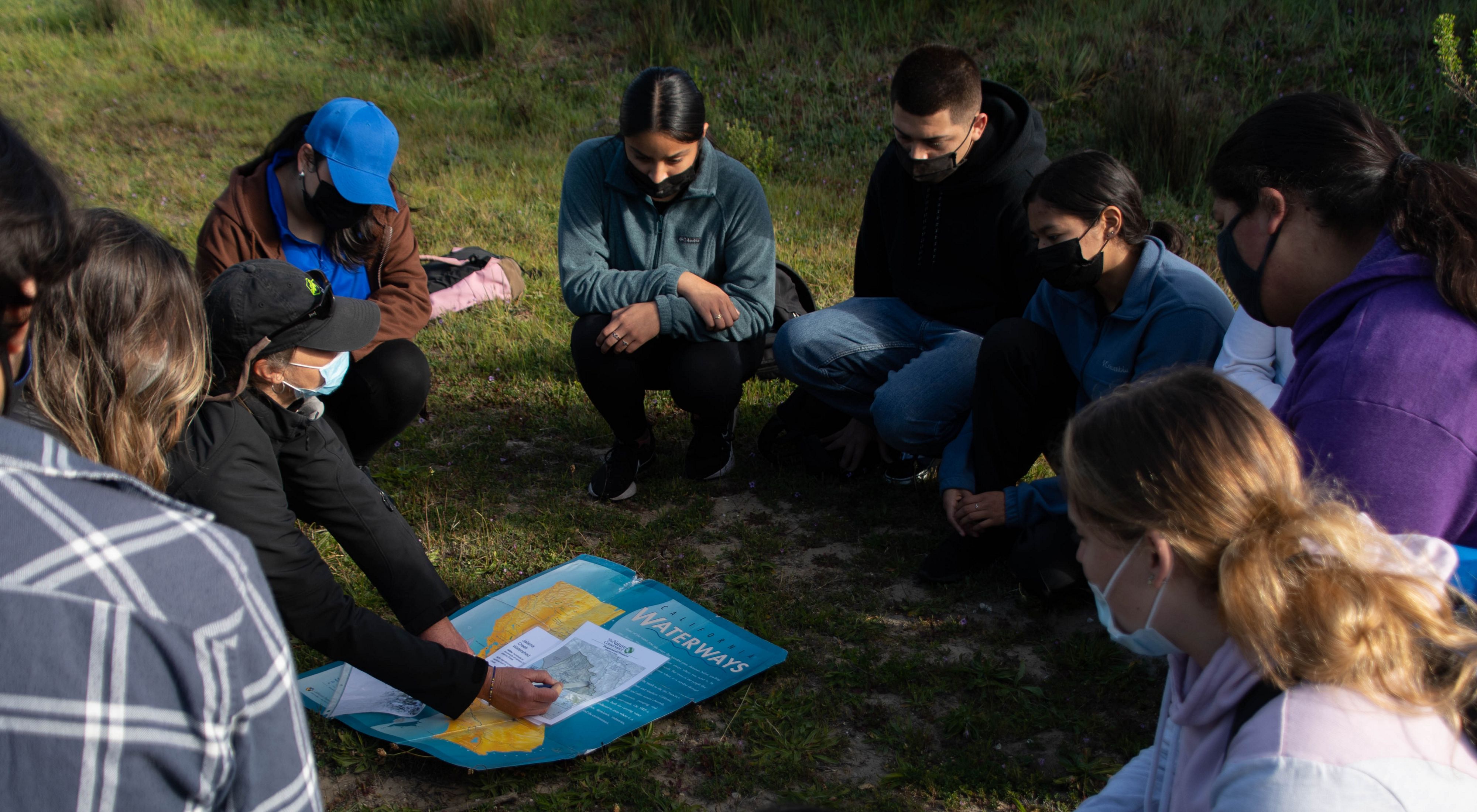 Group of people sitting in a circle in the grass looking down at a map.