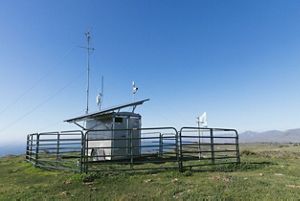 Trailer with an antenna and solar panels attached, surrounded by fencing in a grassy field with ocean in the background.