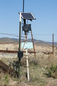 Pole-mounted camera with a solar panel in front of a barbed-wire fence.