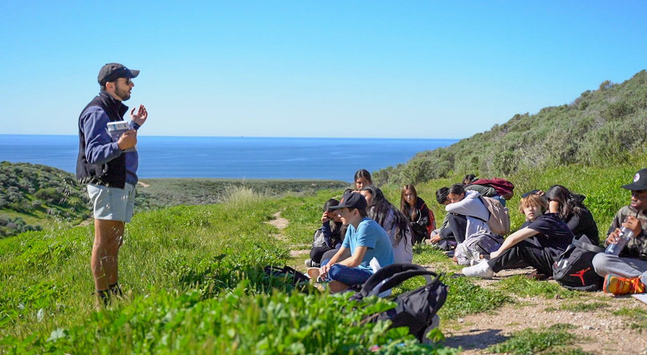 A man stands and speaks in front of a group of sitting students in a grassy meadow with ocean in the background.
