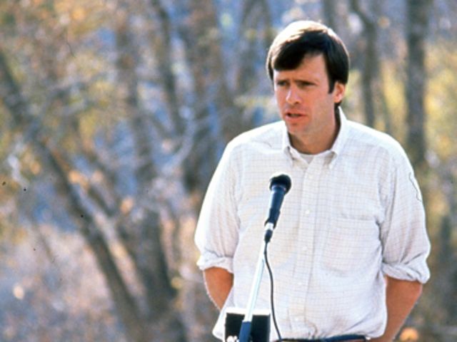 Dave Livermore speaking into a microphone from podium outdoors with tree in the background.