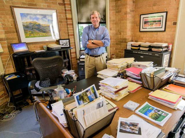 Dave Livermore standing by office desk with piles of papers and magazines.