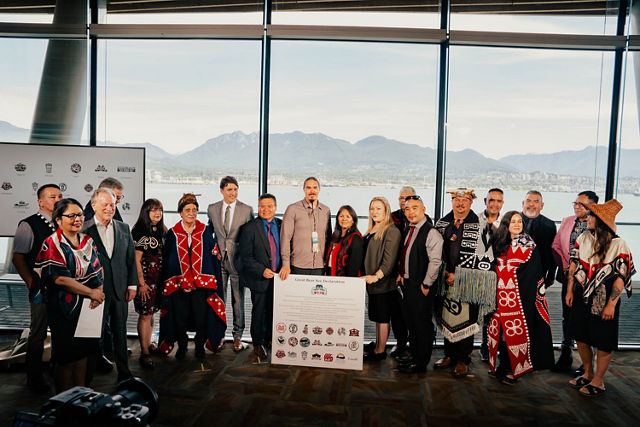 A group of Indigenous leaders wearing a range of formal wear and traditional regalia stand with government officials including justin trudeau, with mountains and a bay in the background