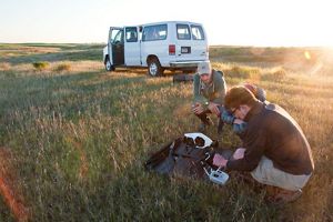 Three students crouch in a field and prepare a drone for a research flight. A white van with the driver's side door open is in the background.