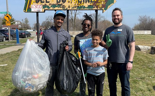 Three adults and a child stand in front of an old sign shaped like an arrow, and one adult is holding two big garbage bags filled with trash.