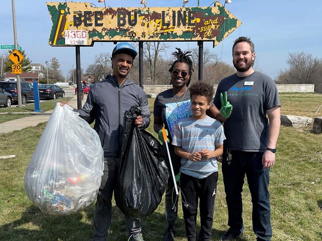 Three adults and a child stand in front of an old sign shaped like an arrow, and one adult is holding two big garbage bags filled with trash.