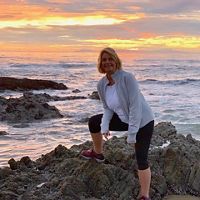 Woman wearing black leggings, white t-shirt and gray jacket stands near an ocean cove with a sunset in the background.