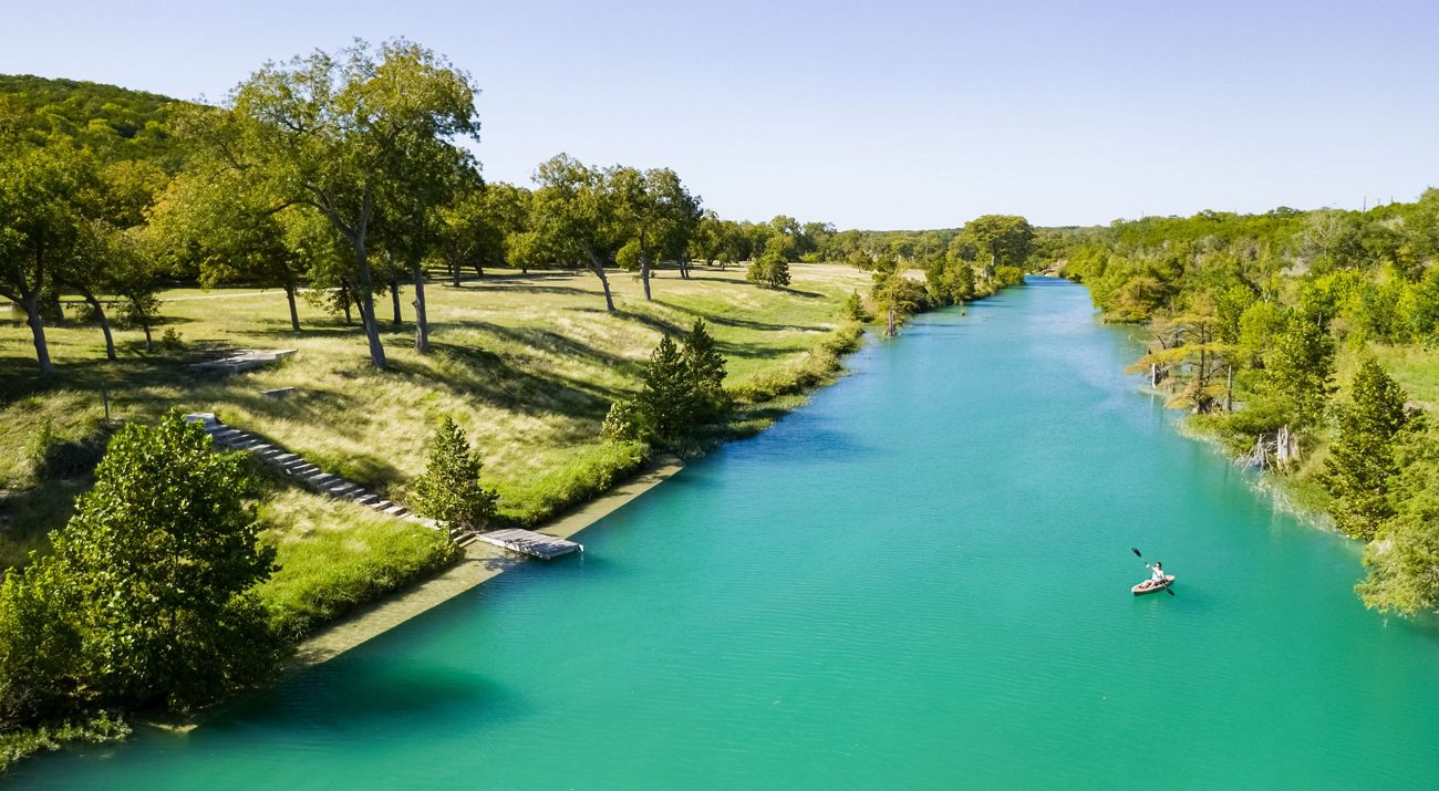 A person kayaks along a wide, blue river that flows between tree covered banks.