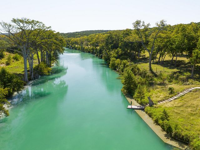 A person stands on a dock overlooking a bright blue river surrounded by green grass.