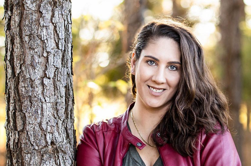Elizabeth Carter headshot. A smiling, dark haired woman wearing a gray top and red leather jacket leans again a tree.