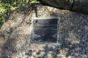A weathered black plaque with raised bluish lettering, mounted to a granite boulder.