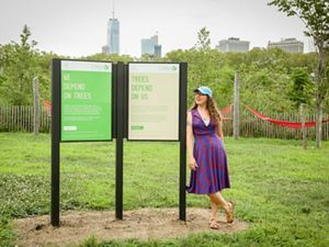 Emily Nobel Maxwell, wearing a purple and red dress and blue TNC baseball cap, leans against a signpost at Governors Island with TNC messaging on two posters. The NYC skyline is in view in the back.