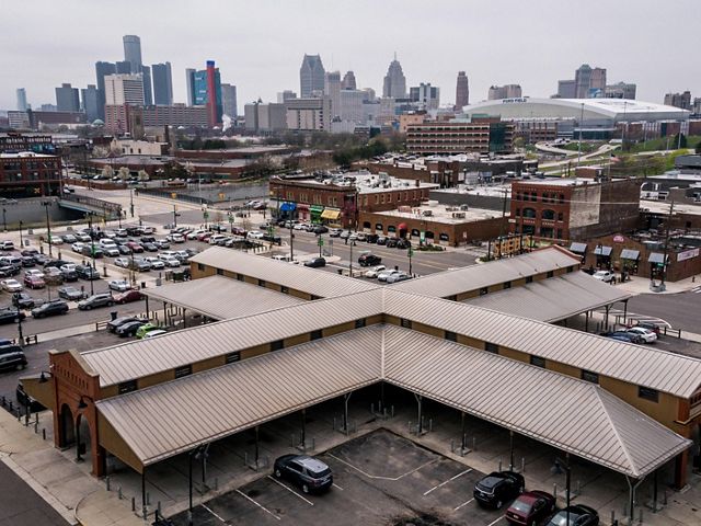 Aerial view of an x-shaped roof over an open-air market surrounded by parking, with the city skyline in the background.