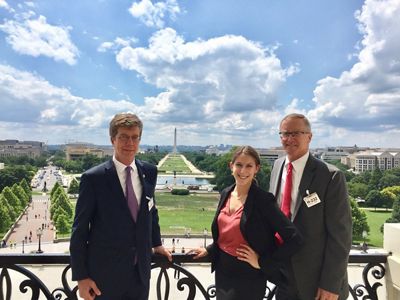Three people in business attire pose on a balcony with the National Mall and Washington Monument in the background.