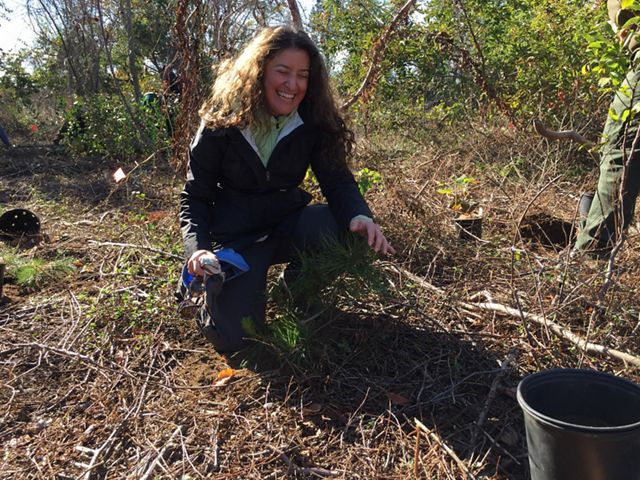 Emily Nobel Maxwell crouches over tree branches and debris onlooking and smiling to the right side of the frame, with a tree plant bucket in view. Green trees are in background.