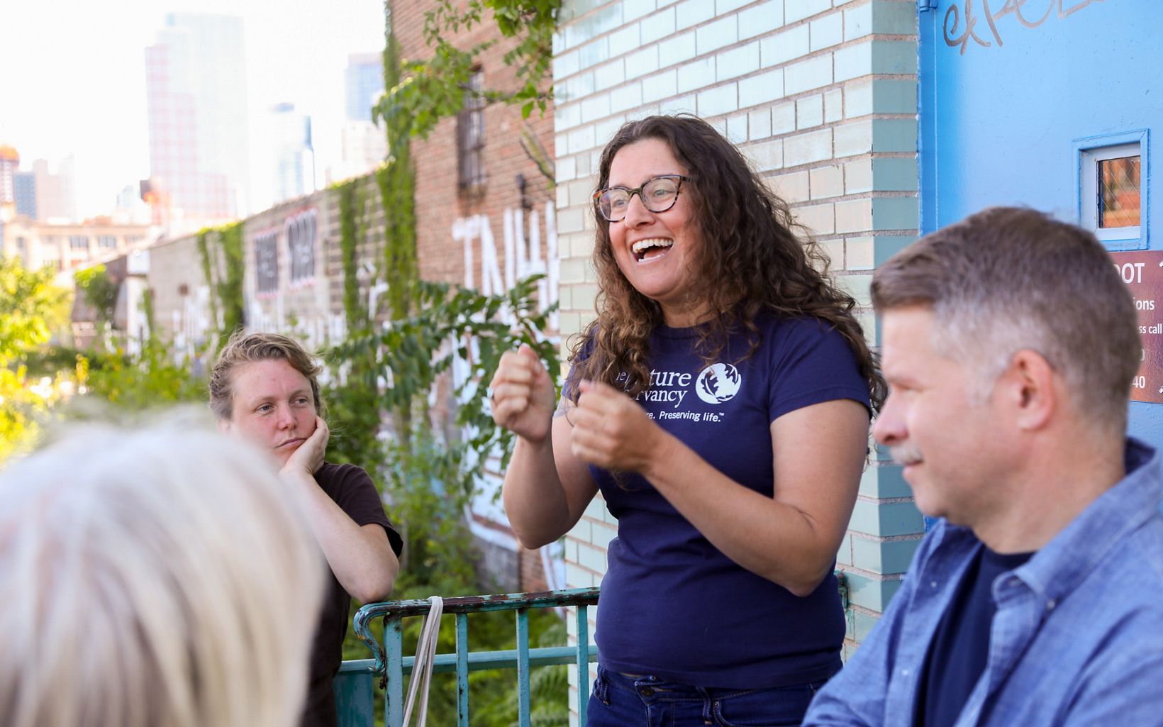 A woman wearing a blue t-shirt with The Nature Conservancy name and logo on it talks with a crowd. The woman is at the right side of the image, has brown curly hair, and wears glasses.