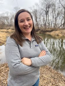 Emily Zimmerman wears a grey sweatshirt and stands outside near a body of water in the fall. 