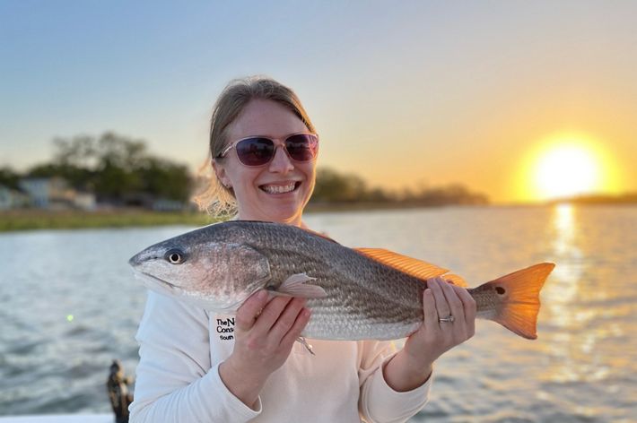 The sun sets behind a woman holding a fish.