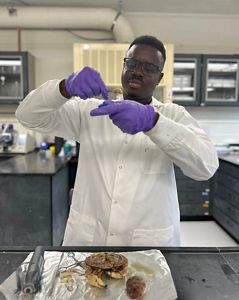 A young scientist dressed in a white lab coat and purple latex gloves stands behind a table preparing a tissue sample taken from a Jonah crab on a tray in front of him.