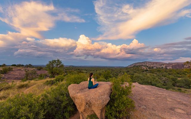 A woman sits atop a large, orange boulder looking out at a rocky dome dotted with green trees.
