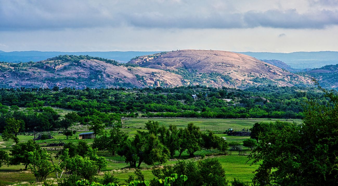 A dome of pink rock towers over green fields with trees.