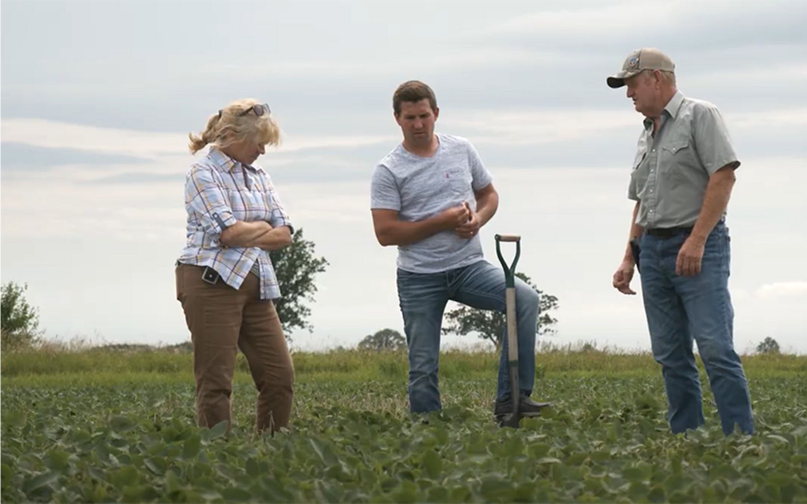 Three people stand in an agriculture field with a shovel as they discuss soil and nutrient management. 