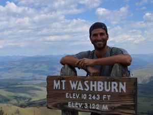 Man standing on a mountain behind a "Mt Washburn" sign