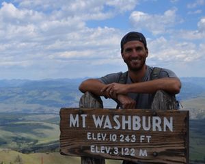 Man standing on a mountain behind a "Mt Washburn" sign