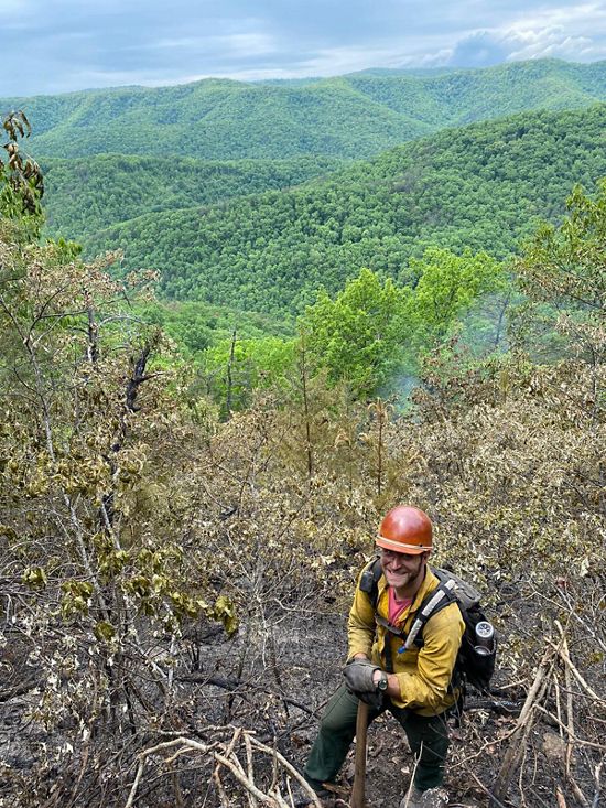 Candid photo of Eric Homan standing on a fire line during a controlled burn with rolling mountain ridges behind him.