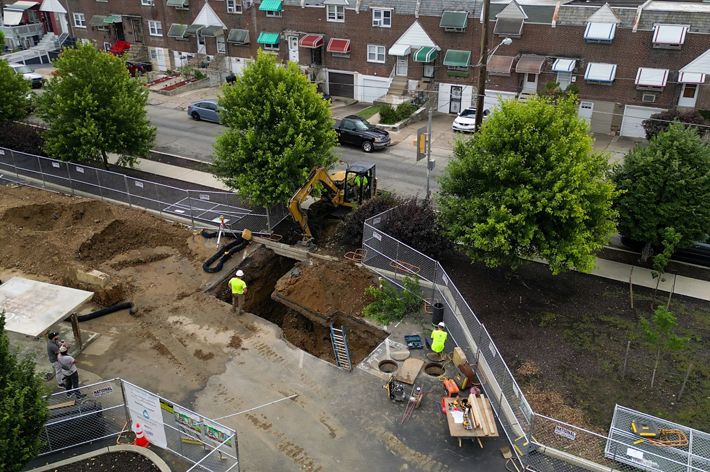 A drone photo looking down onto a parking lot with large yellow construction vehicles and workers digging a hole into brown dirt.