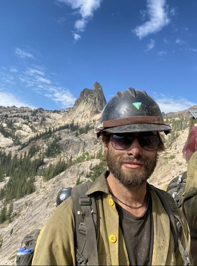 Ethan is smiling while in prescribed fire gear with mountains in the backdrop.
