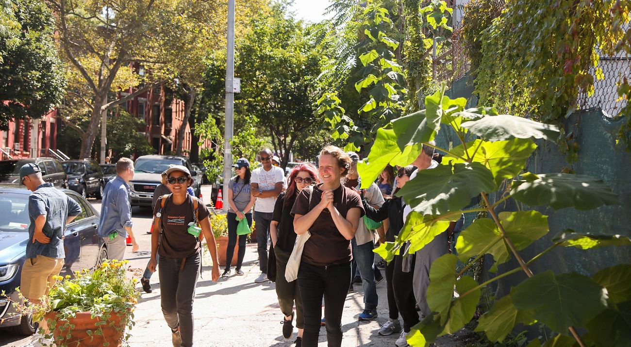 A group of nine people in view walking down a city sidewalk with a fence with greenery at right and a street with parked cars on the left. Brownstone buildings are seen in the background.