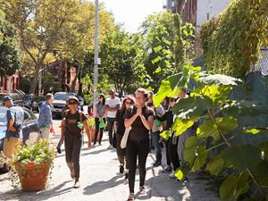 A group of nine people in view smiling and walking down a city sidewalk with a fence with greenery at right and a street with parked cars on the left. Brownstone buildings are seen in the background. 