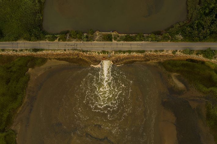a cracked road stretching over a creek is seen from above with water spilling out of a narrow hole on one side