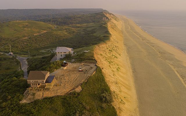 a steep sand bluff is seen from above with two houses near the edge. One house sits farther back from the edge with construction equipment nearby