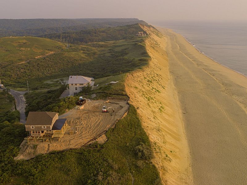 a steep sand bluff is seen from above with two houses near the edge. One house sits farther back from the edge with construction equipment nearby