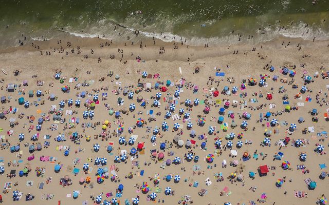 Seen from above: dozens of beach umbrellas and towels dots a beach with swimmers standing on the edge of the water