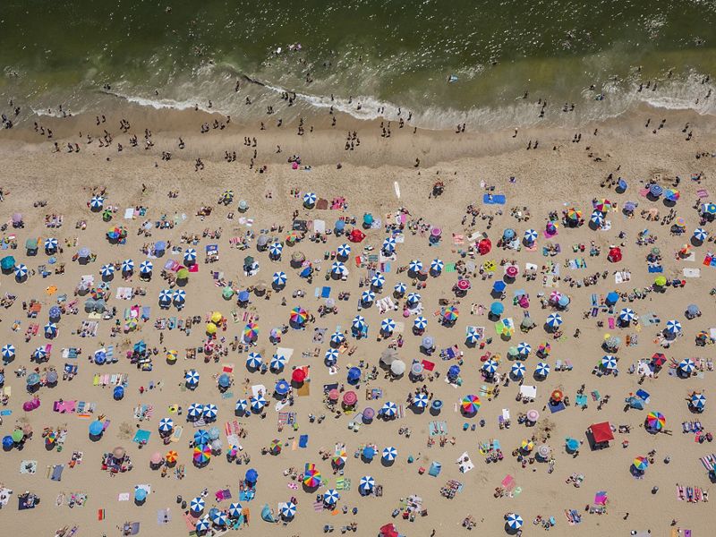 Seen from above: dozens of beach umbrellas and towels dots a beach with swimmers standing on the edge of the water