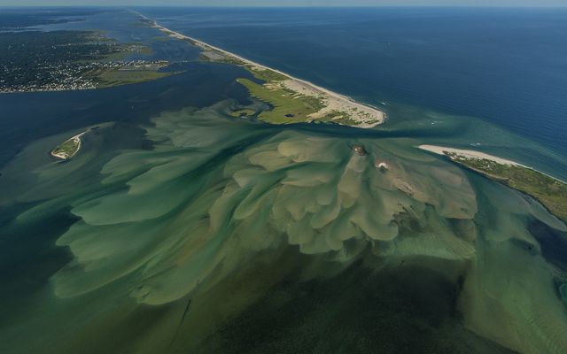 seen from above: a narrow stretch of land surrounded by blue ocean water on both sides is shown with a cut in the middle of the land where the water has pushed through, mud/sand visible on the water