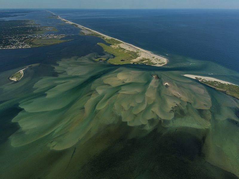 seen from above: a narrow stretch of land surrounded by blue ocean water on both sides is shown with a cut in the middle of the land where the water has pushed through, mud/sand visible on the water