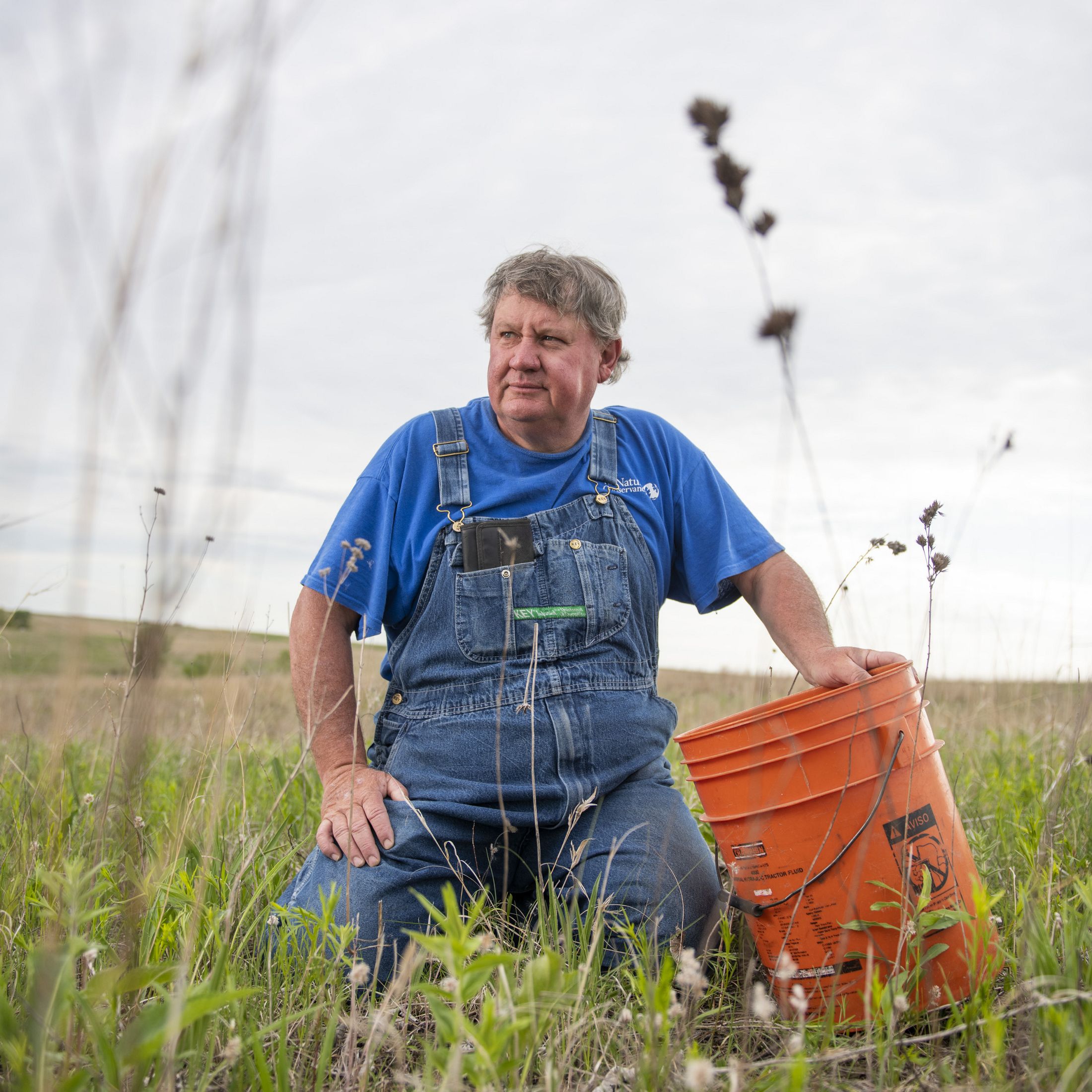portrait of man in overalls sitting in a field with an orange utility bucket