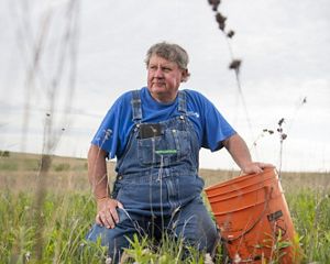 portrait of man in overalls sitting in a field with an orange utility bucket